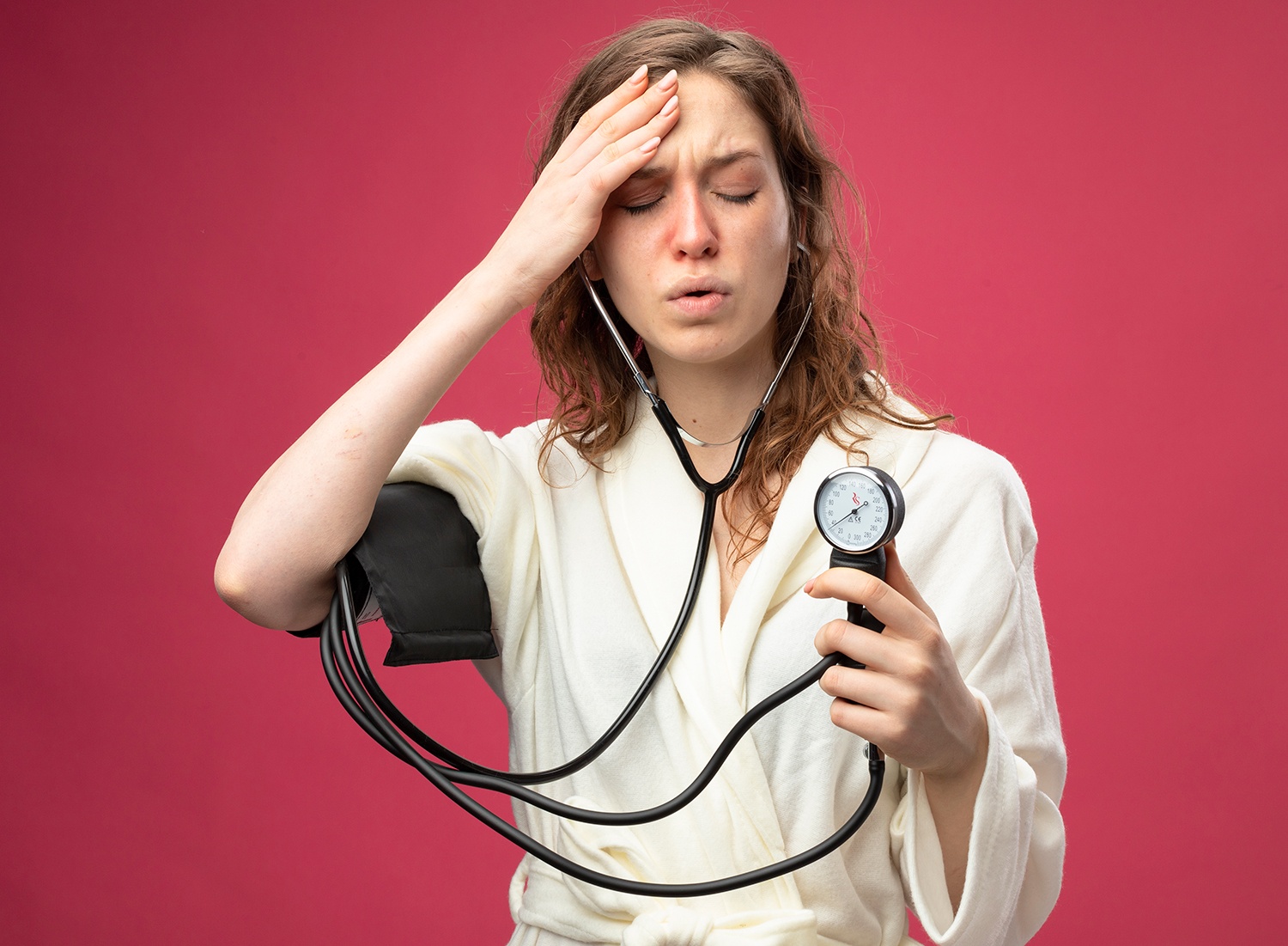weak young ill girl with closed eyes wearing white robe measuring her own pressure with sphygmomanometer putting hand on forehead isolated on pink background